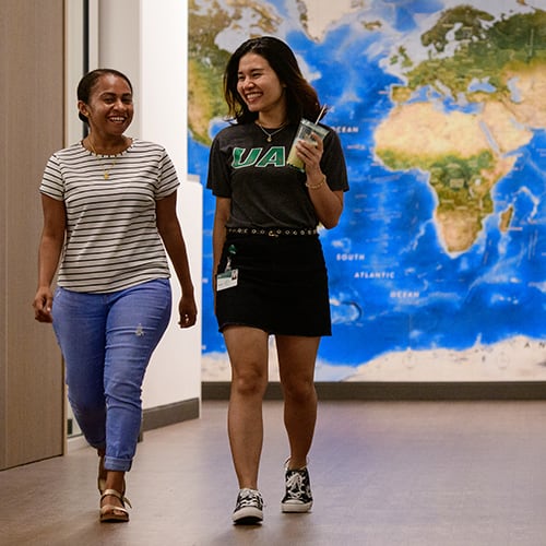 Two students walking down the hall with a map of the world behind them.