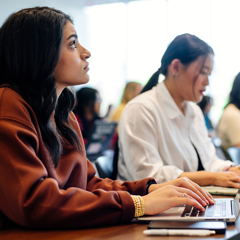 Student with long dark hair in class looking up at professor with fingers on laptop.