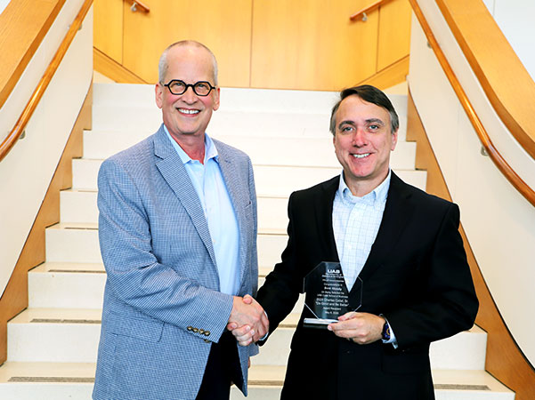 two people in front of a staircase shaking hands and smiling. One has a crystal award in hand.