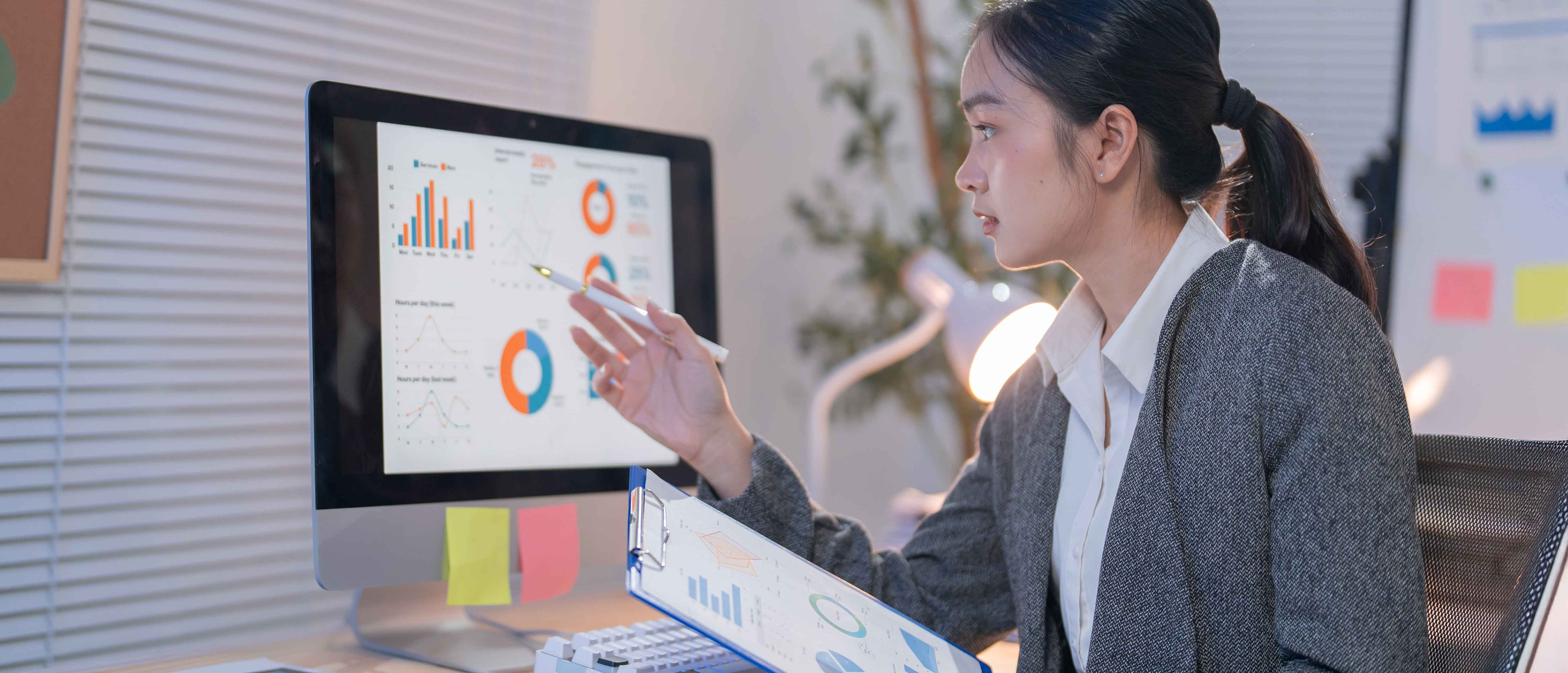 Person analyzing data dashboards—pointing a stylus at bar and pie charts on a desktop monitor while holding a clipboard of graphs in a modern office.