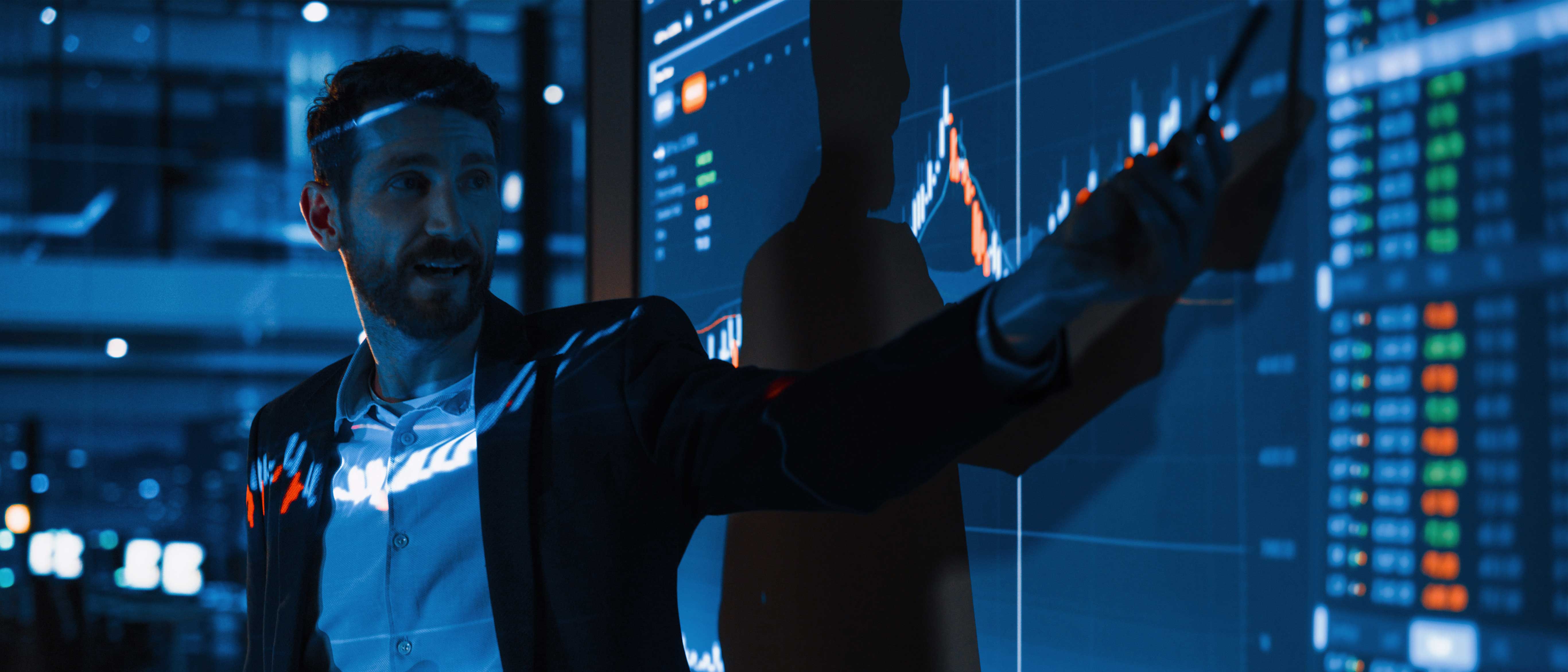 Man in business attire pointing at a large digital screen displaying financial charts and stock market data in a dimly lit office environment.