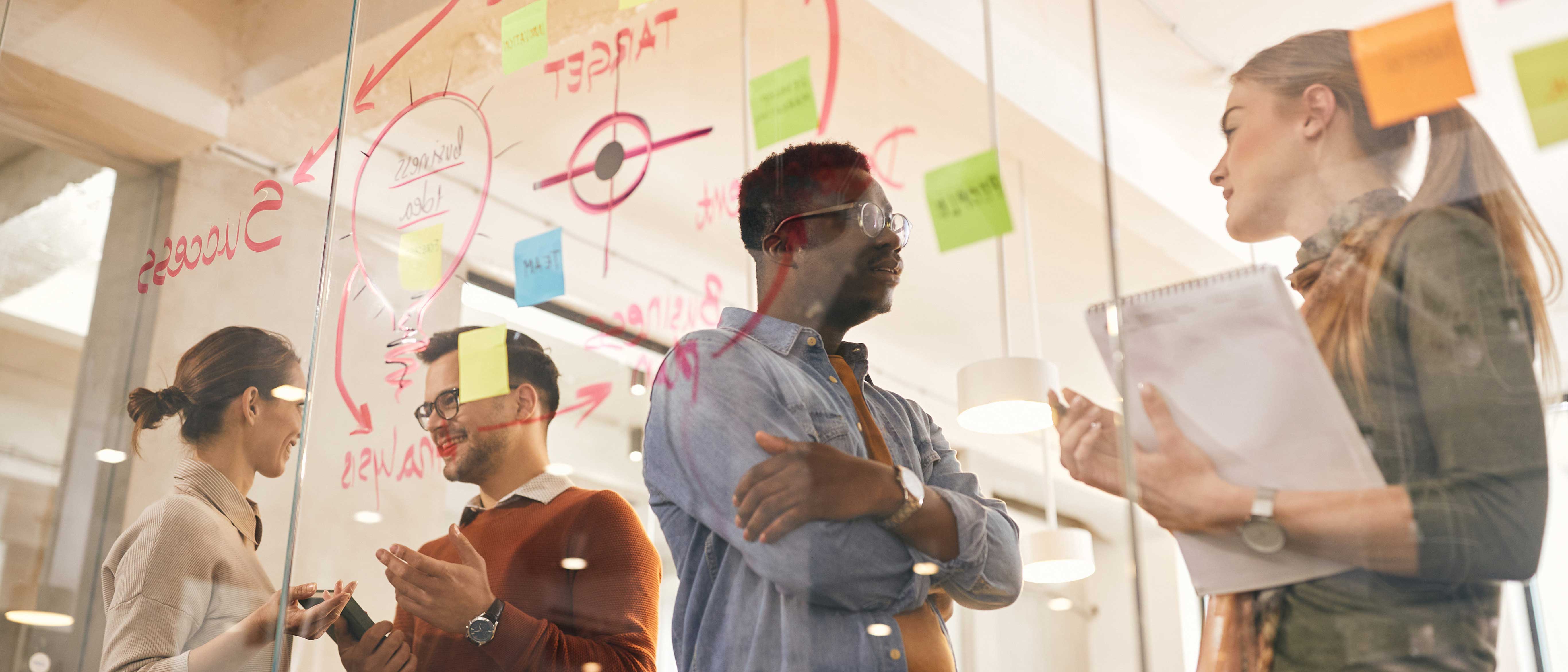 Four people are having a discussion behind a glass wall covered with red marker drawings and colorful sticky notes in an office.