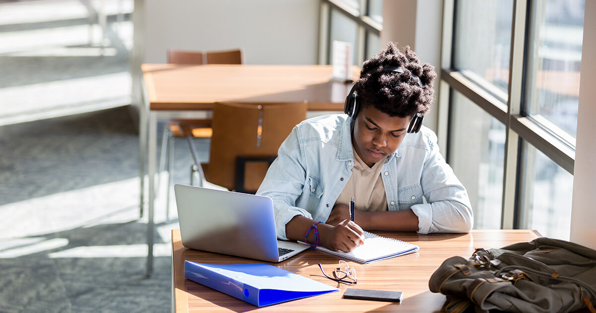 A student with his headphones on writing in a notebook, his laptop open next to him. 