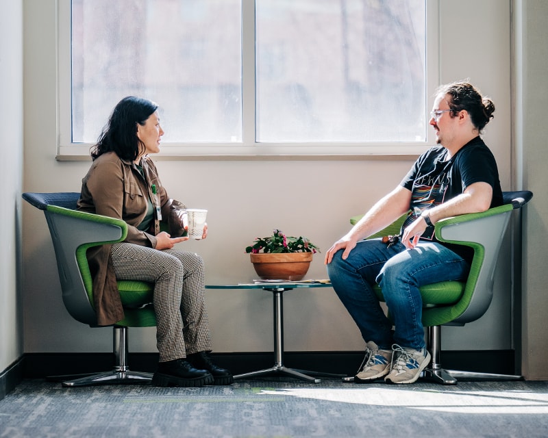 Student talking with faculty member, both seated in front of a large window in University Hall. 