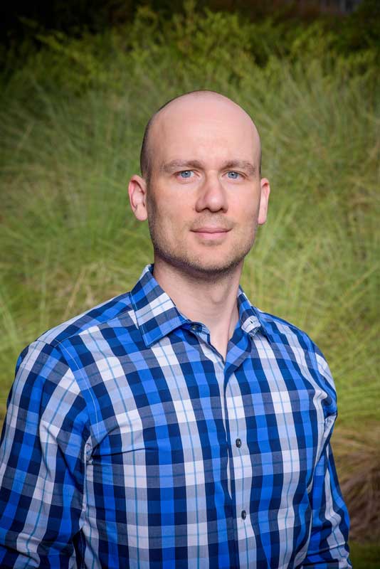 McCain standing in front of a large grassy plant in the evening wearing a blue plaid shirt.