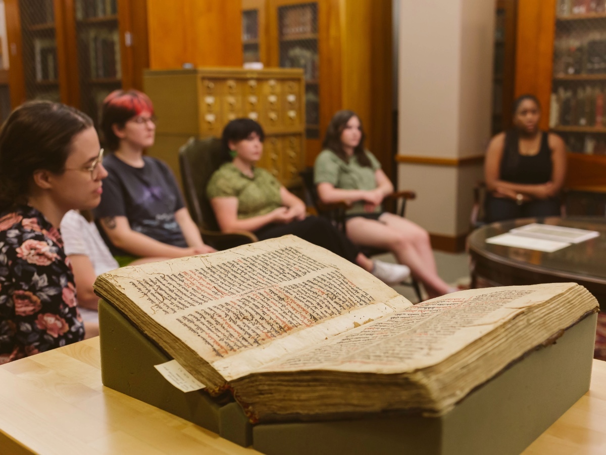 A religious book sits open in the foreground as students discuss the text in the background
