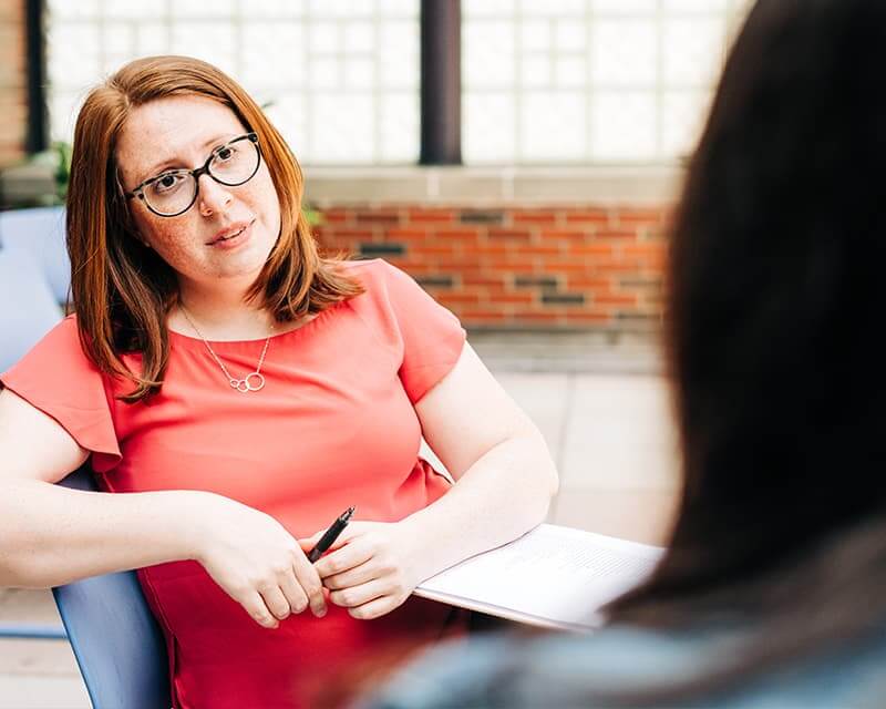 Red-haired woman in a coral shirt and glasses with a notebook, listening to person in foreground.