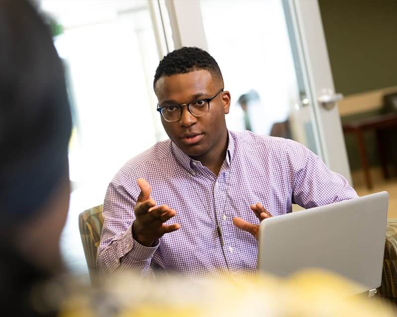 Black male in glasses and a purple checkered shirt, talking to someone hazy in foreground.