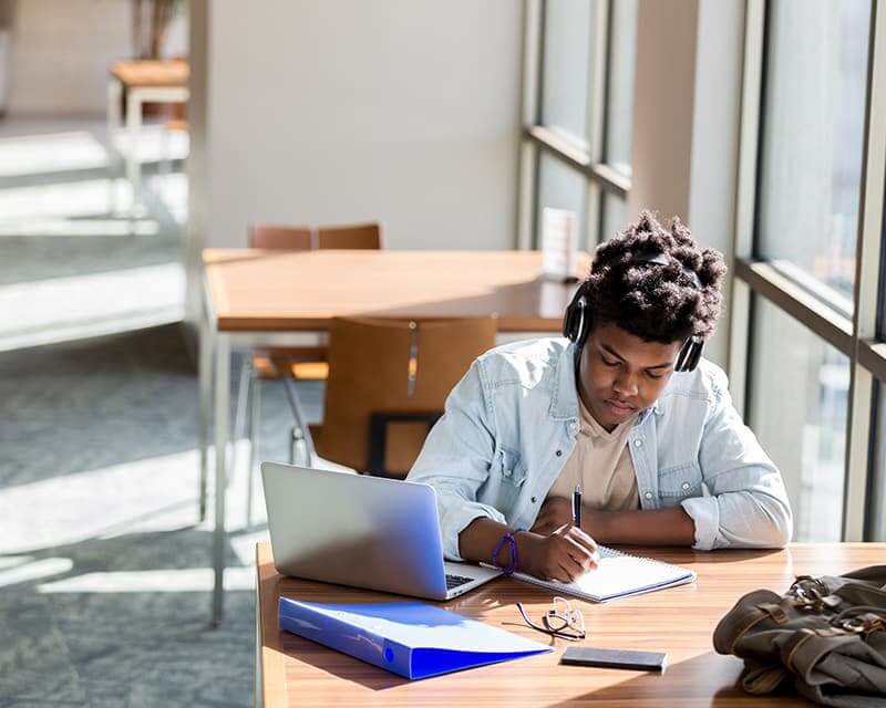 Black male student writing in a notebook at a sunny table next to a window. 