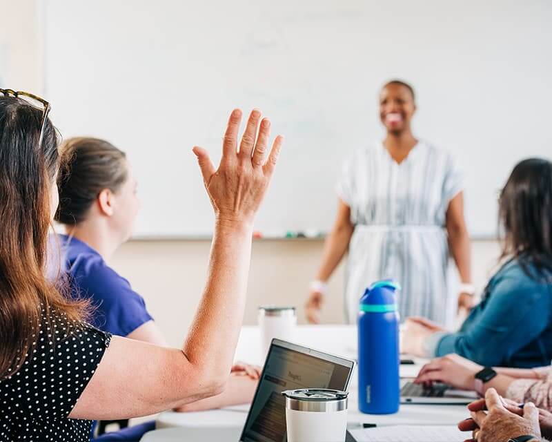 Students at a table, a woman raising her hand in foreground, a black woman teacher in background.