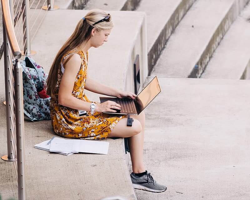 Female student with long blond hair studies outside, wearing a yellow sundress and gray sneakers. 