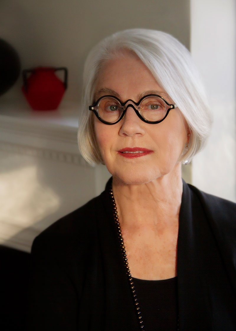A middle-aged woman with a sleek white bob, round black glasses, black top, and shiny black bead necklace.