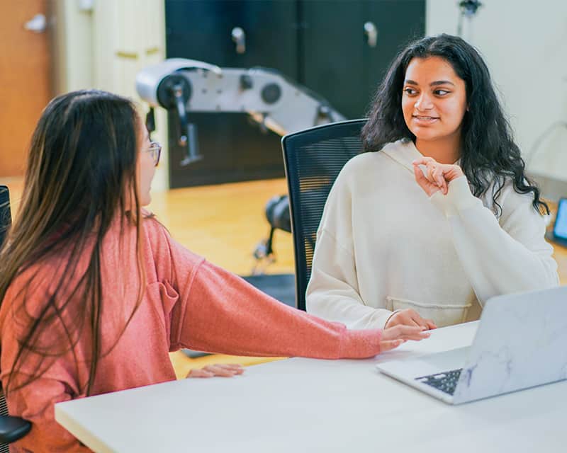 Two young women with long dark hair talk at a table. 