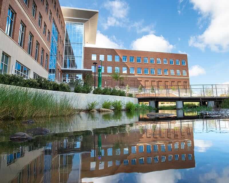 A UAB building reflecting in a lake, with blue sky and white clouds in the background. 