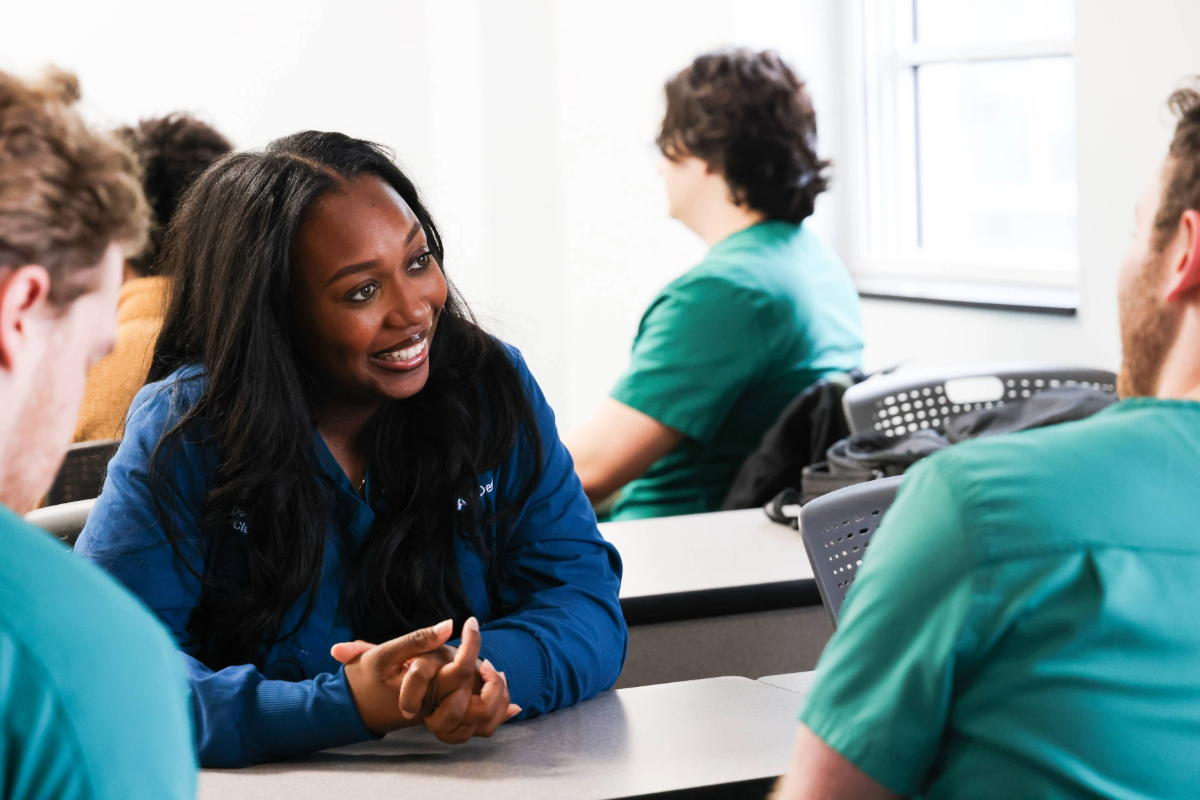 Woman in blue scrubs smiling at person in green scrubs across from her over a table in a classroom setting