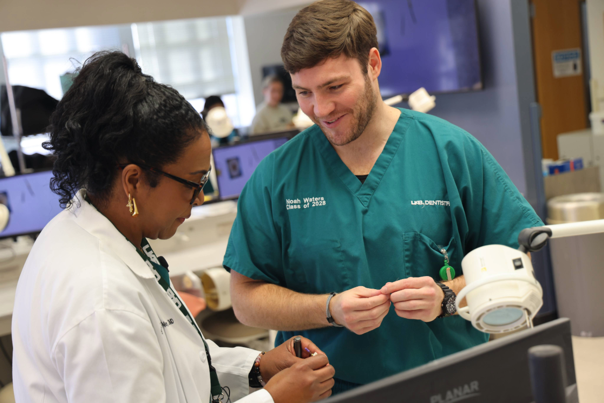 Woman with black hair and white coat (Dr. Coble) assisting a man in green scrubs (dental student) in the preclinical dentistry lab