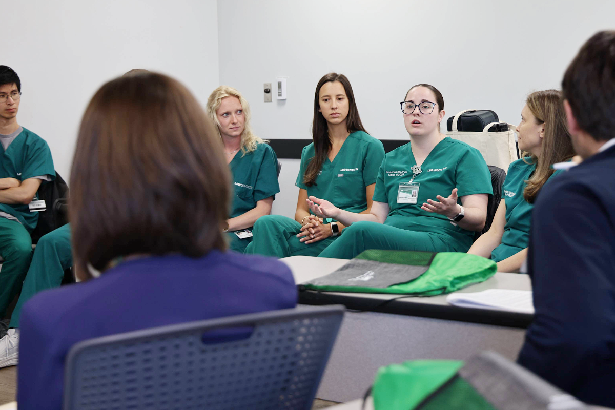 Image of several people in green scrubs and one woman talking in a group meeting to two people in blue suits in a classroom setting