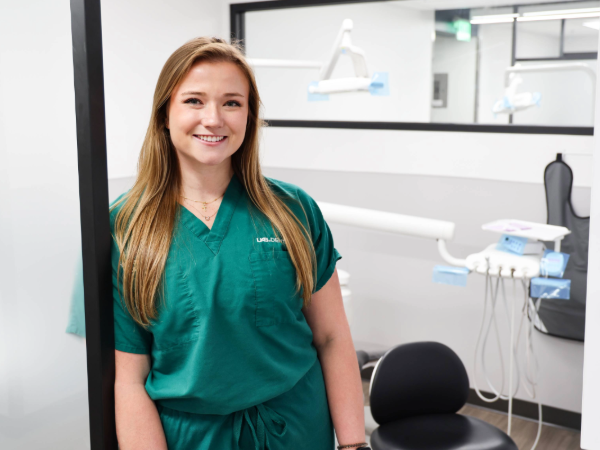 Smiling dental student wearing green scrubs in a clinical training room, with dental equipment and chair visible in the background.