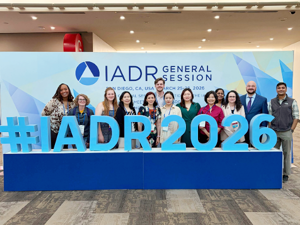 Image of group of people standing behind a large sign that reads, " #IADR2026"