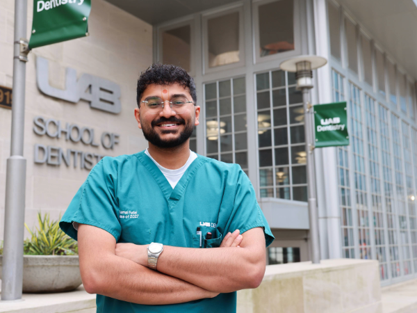 Male dental student (Kamal Patel) smiling with arms crossed wearing green scrubs in front of the UAB School of Dentistry building