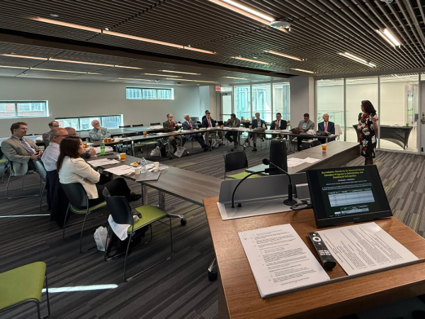 Wide-shot image of people in professional attire in a meeting room sitting at a conference table listening to a woman speaker standing to the side