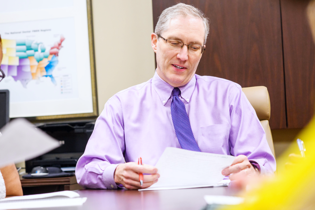 Man in a purple shirt and tie reviewing a document at an office table during a meeting.