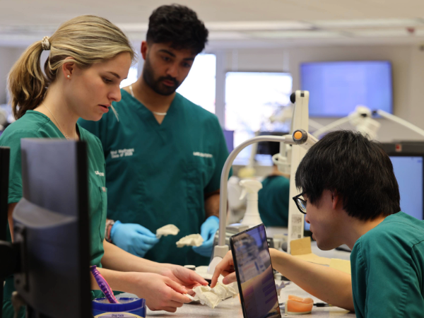 Two dental students in green scrubs holding teeth models in a teaching moment with a sitting faculty member in green scrubs