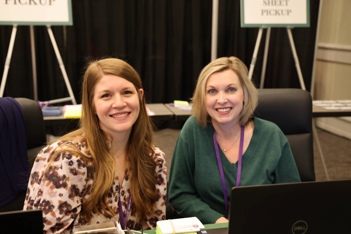 Two women in professional attire smiling for a photo at a registration desk at a conference
