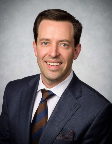 SOD Alumni Kyle Trammell - White man with brown hair wearing a blue suit smiling in front of grey background