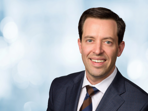 SOD Alumni Kyle Trammell - White man with brown hair wearing a blue suit smiling in front of blue and white background