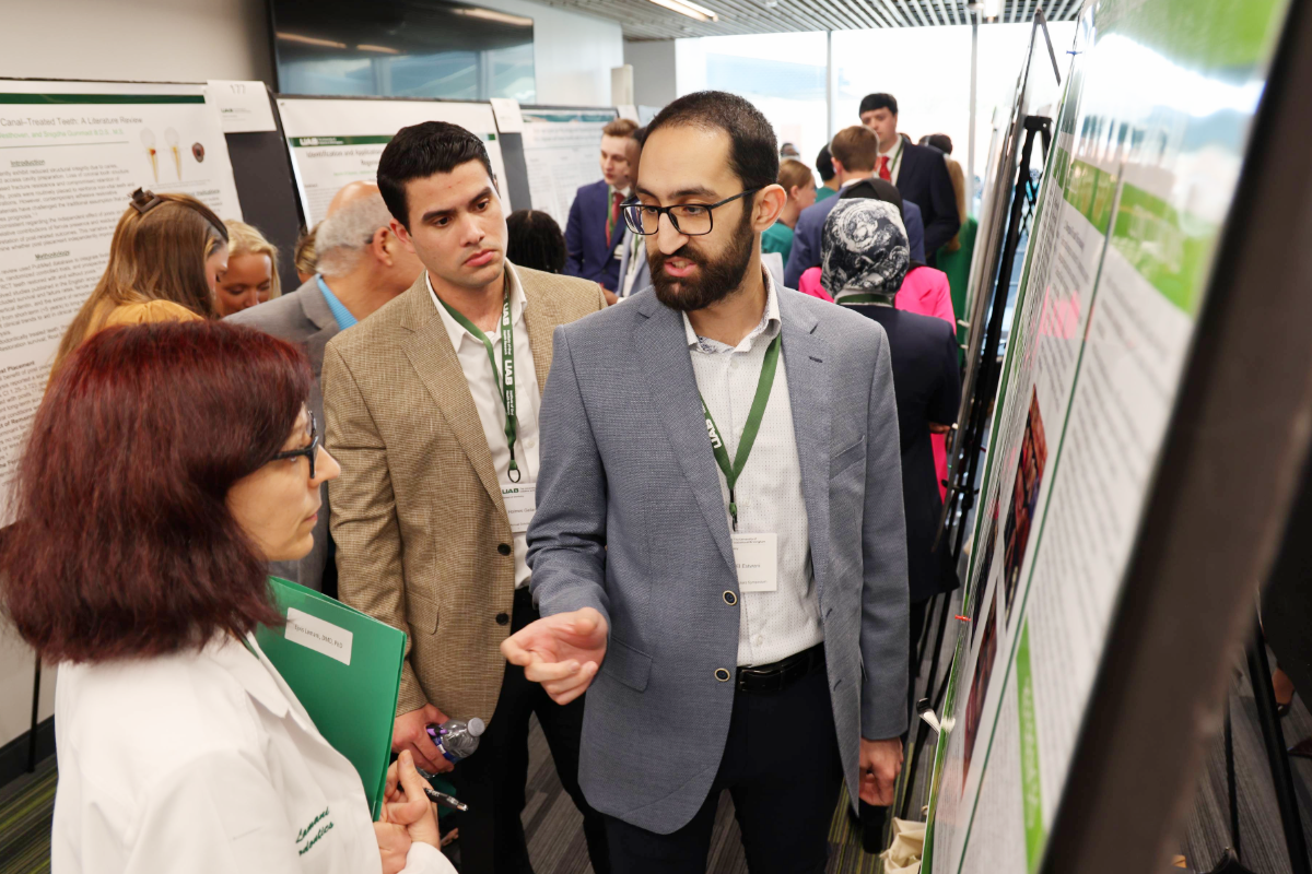 Male dental student in suit talking to female faculty member holding green folder in front of research poster among a crowd of other dental students and faculty