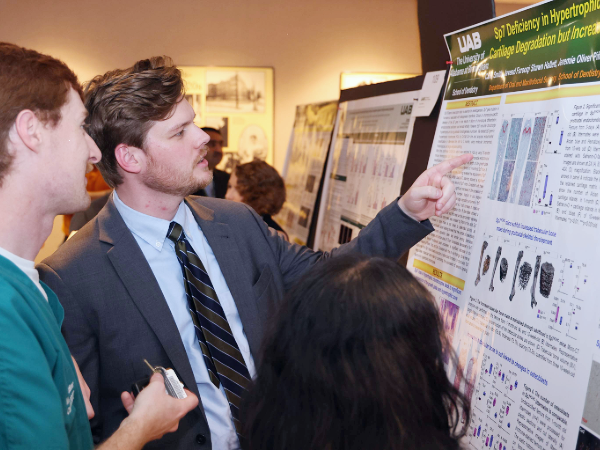 Male wearing suit pointing at research poster next to male student in green scrubs