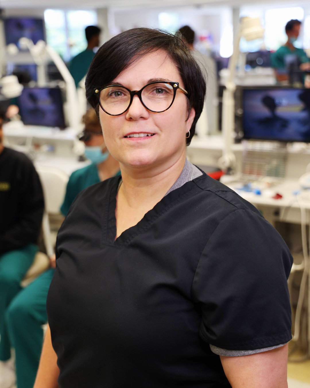 Woman with black hair, glasses and scrubs standing in a preclinical dentistry lab that seems to be busy
