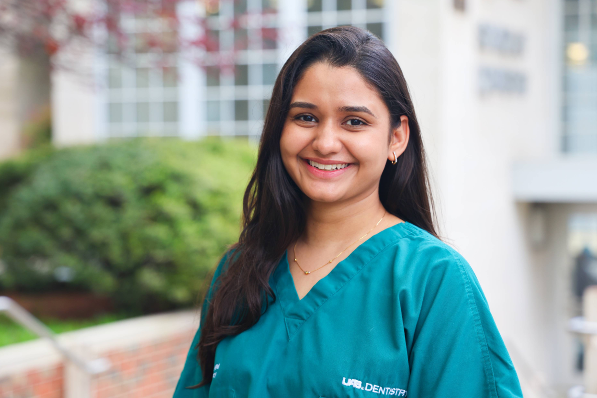 Woman with brown hair and green scrubs smiling with a blurred UAB School of Dentistry building in the background