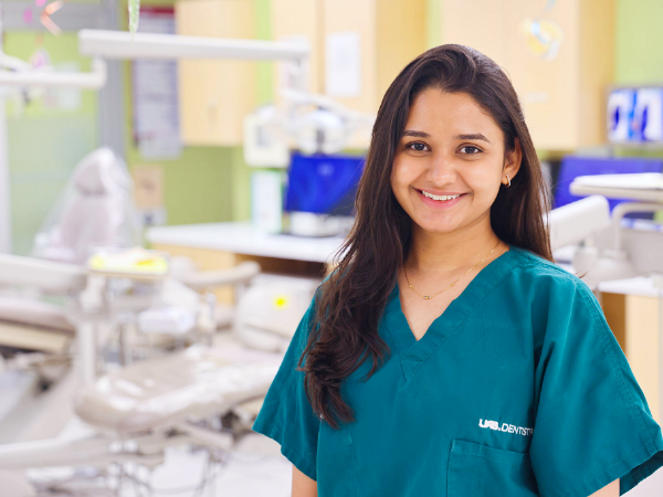 Woman with brown hair and green scrubs smiling with a blurred dental clinic setting in the background