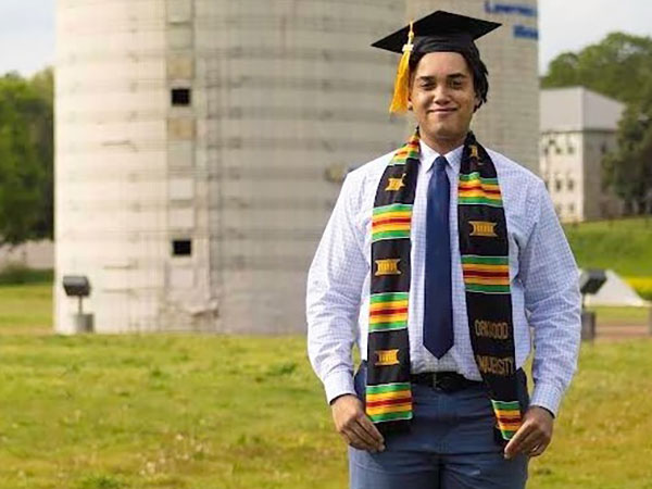 Emmanuel Williams standing in front of a grain silo wearing graduation cap and regalia. 