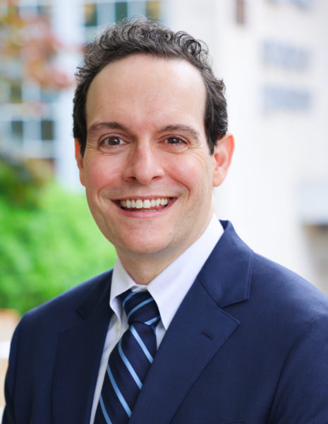 Man in professional attire smiling outside building with greenery