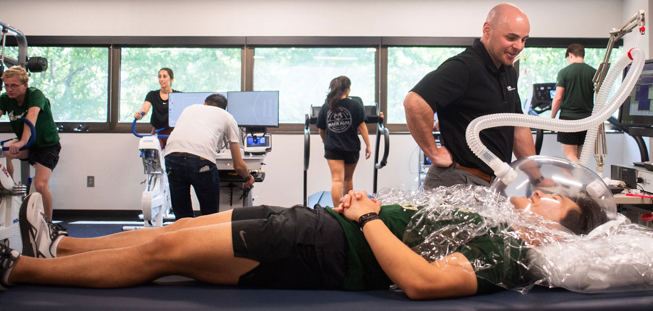 In a gym setting, a physical therapist works with a woman lying down with an elaborate breathing mechanism on her head and shoulders. 