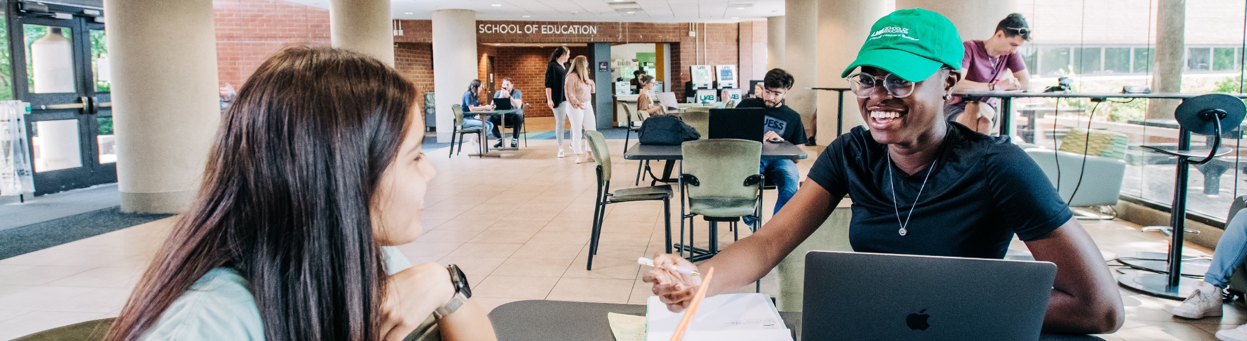 Students sitting in a lounge area in the school, a smiling black female in a green UAB hat in the foreground.