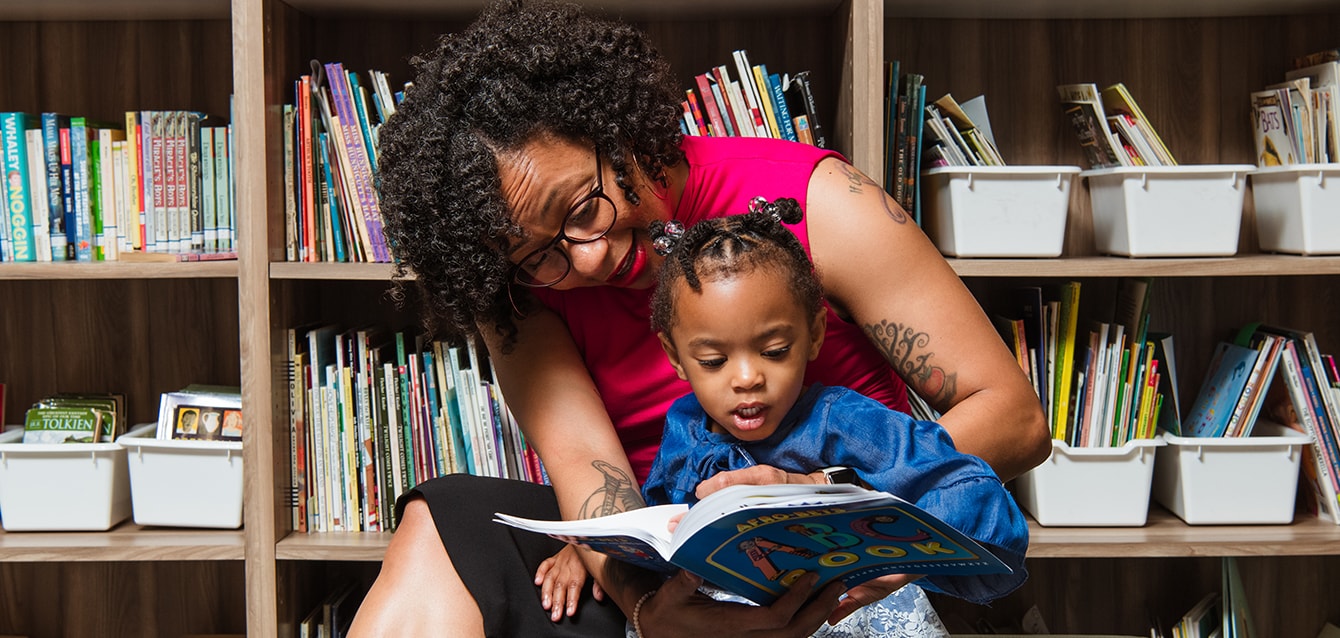 A female teacher with natural hair and tattoos sits in front of a book case reading to a small child. 