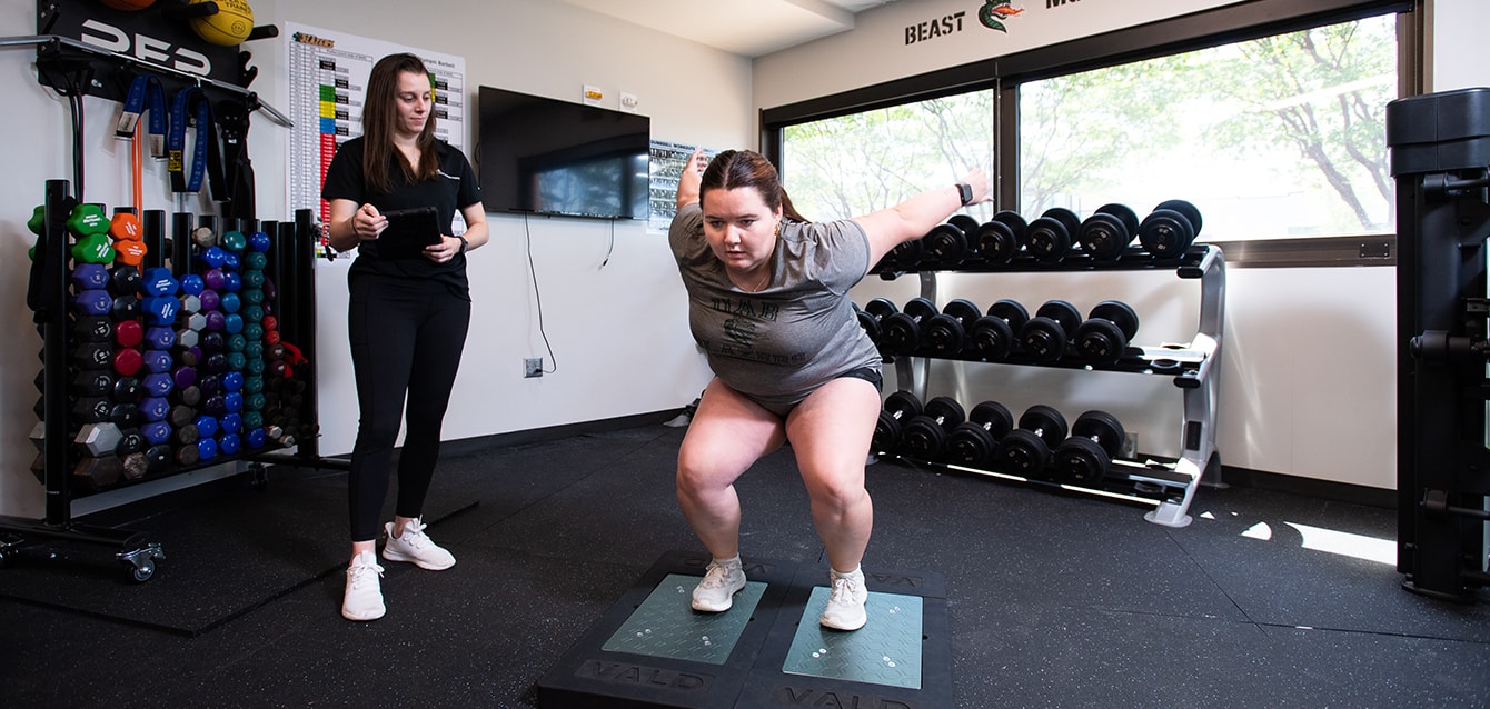 A female trainer in a UAB facility full of hand weights watches a young woman perform a squat. 