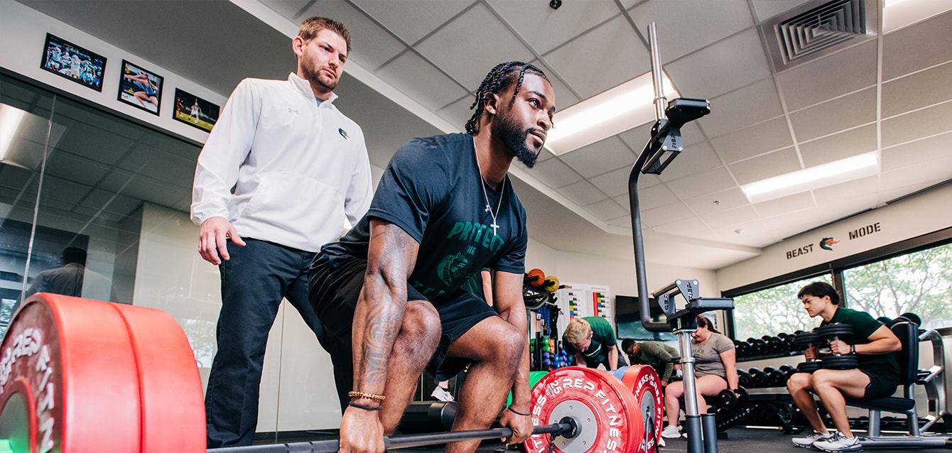 A male trainer watches as a young man squats to lift a heavily weighted barbell. 