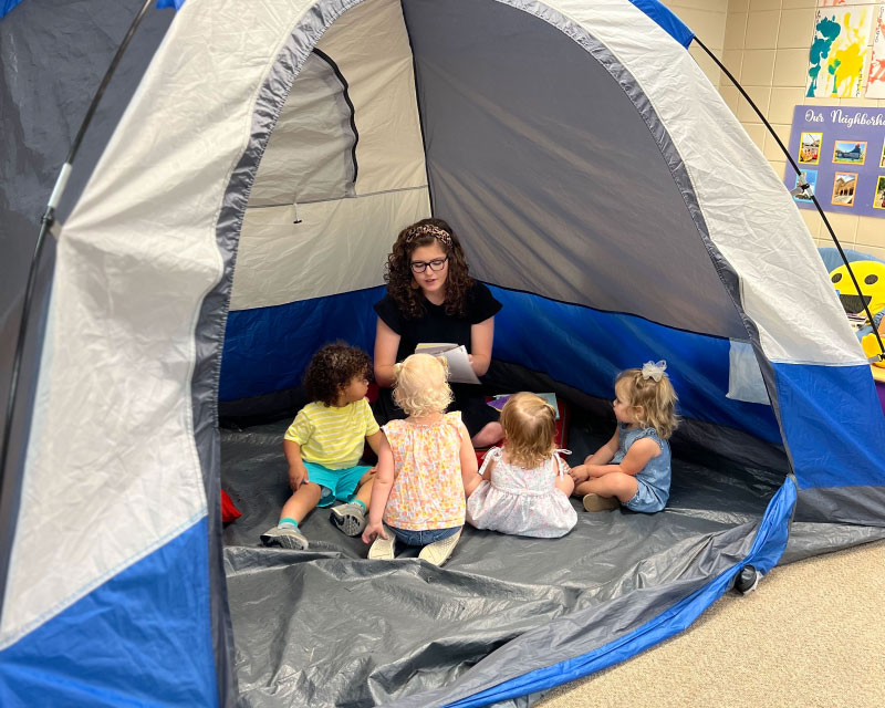 Teacher candidate sits inside a tent that's been set up inside a classroom, reading to preschoolers. 