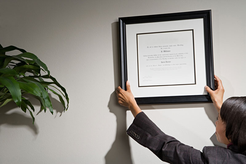 A woman hanging a framed diploma on the wall of an office.