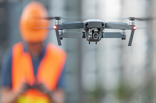 A drone hovers in the foreground, flown by a person in a hardhat who is blurred in the background