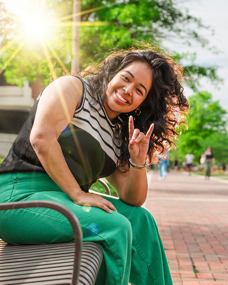 Female student sits on a bench on UAB's campus while making the Blazer sign with her hand.