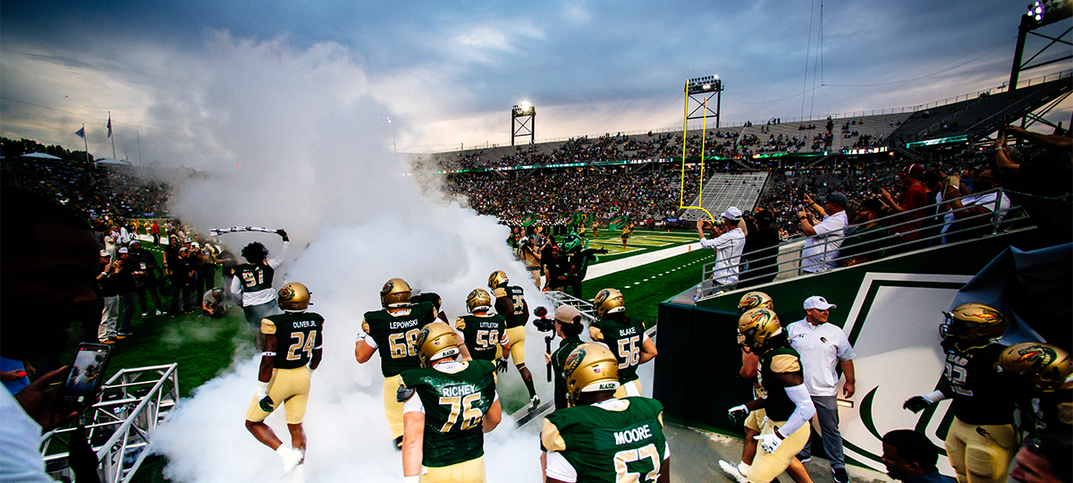 Photo of UAB Blazer football players running through a smoke machine onto the field at twilight.