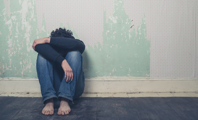 Barefooted depressed man with his head on his knees sitting against a wall.