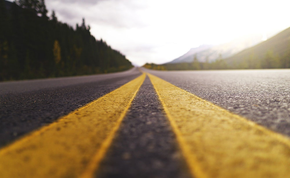 Double yellow line of a road stretching away to the horizon, trees on the left and a mountain range on the right.