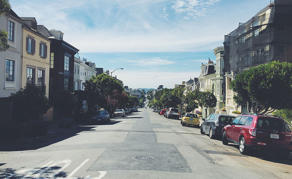 Looking down a patched residential street lined with townhouses, a cloudy blue sky above. 